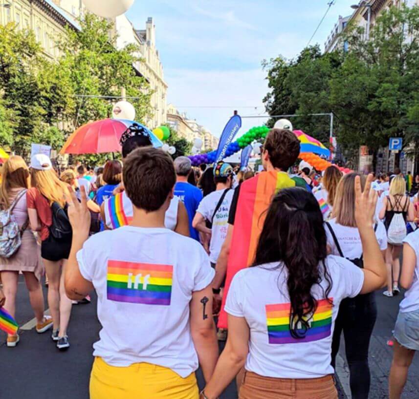 Participants walking in a Pride parade along a city street, surrounded by rainbow flags and balloons. In the foreground, two individuals wearing white T-shirts with rainbow stripes and an IFF logo hold hands, while others nearby carry Pride-themed accessories