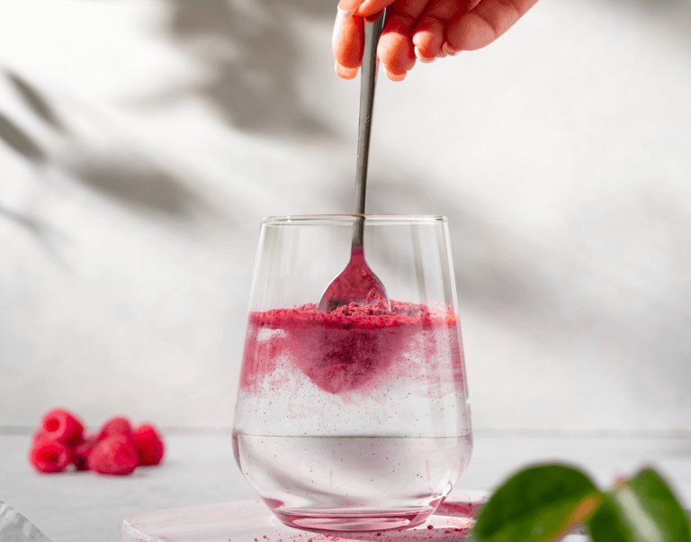 Woman's hands stirring raspberry-flavored powder into her glass of water. Raspberries sitting next to glass