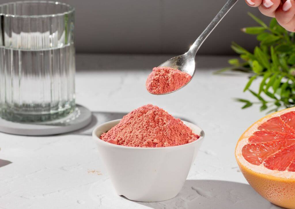 Hand holding a spoon, picking up grapefruit-flavored beverage flavor powder, getting ready to sprinkle into a glass of water. Water and a grapefruit sitting next to cup of flavor