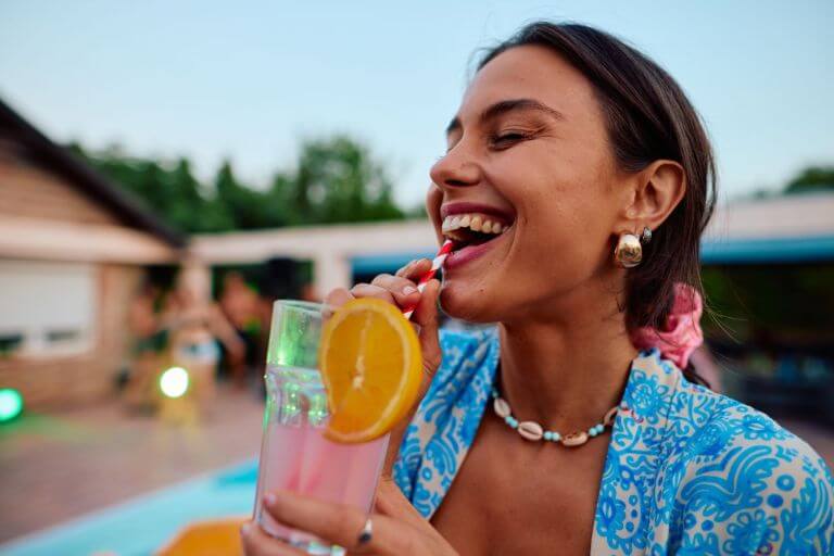 Woman outside at a party, laughing, dressed up and sipping a citrus-flavored beverage with a straw through her teeth.