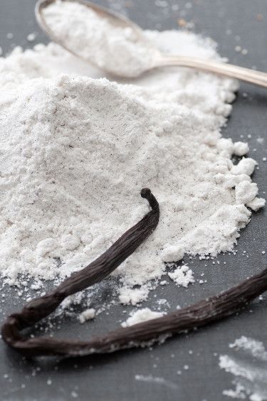 Ground vanilla-flavored powder sitting in a pile on a granite counter with a spoon next to it and two vanilla pods in the front