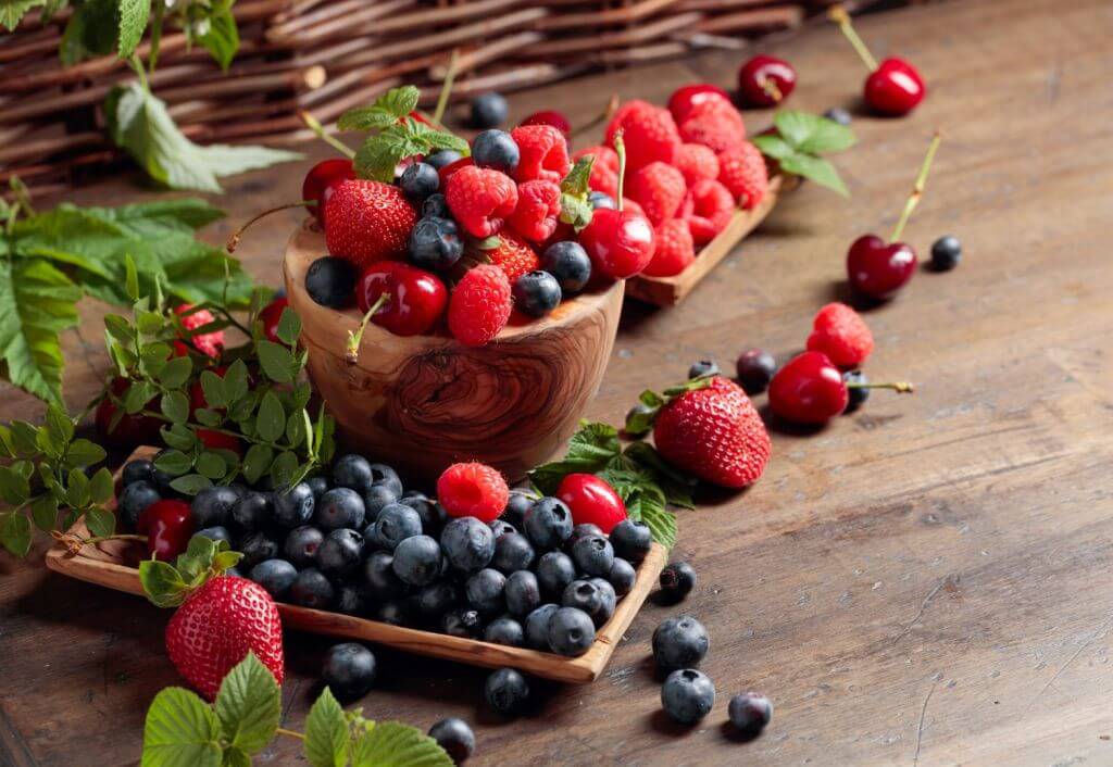 Bowls and trays of multiple types of berries and berry leaves sitting on a wooden table. Side of a brown wicker chair in the background