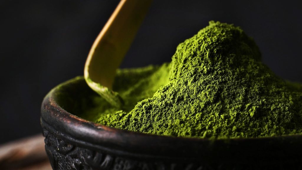 Green matcha powder piled up in a dark bowl with embellishments. A wooden scoop is about to scoop up some of the matcha powder.