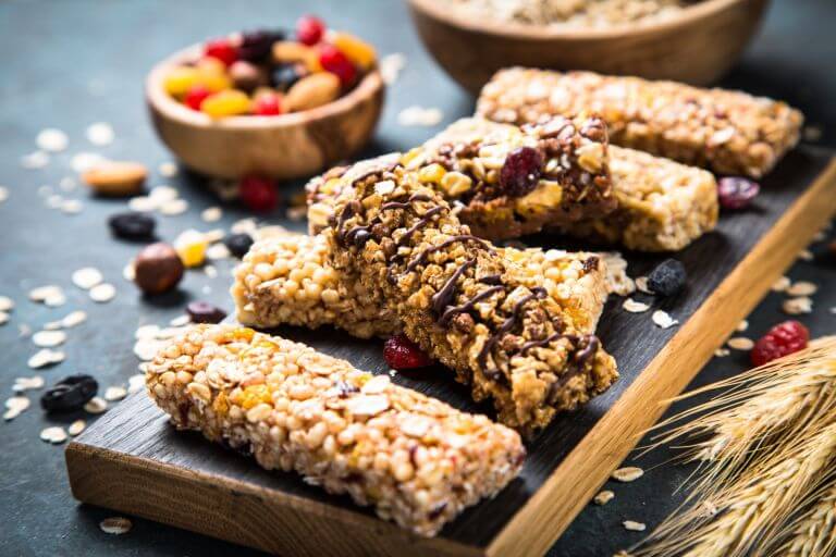 Snack bars all lined up on a counter, some stacked on top of others, with bowls of dried fruits and wheat around the bars