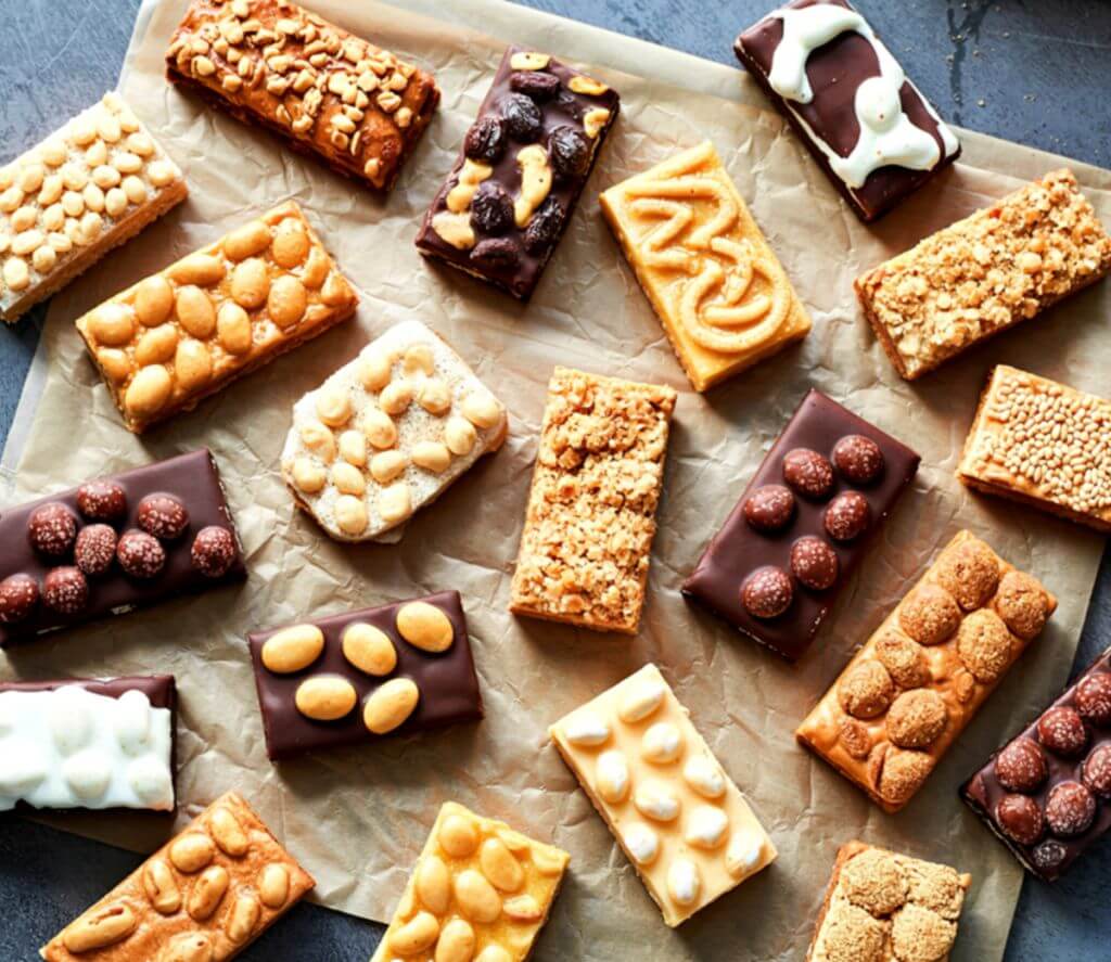 A variety of snack bars, including some with peanuts, some with chocolate and berries, some with white frosting and more, all arranged on unbleached parchment paper and laid out on a counter top