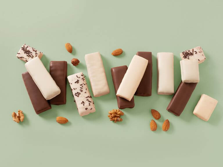Vanilla- and chocolate-coated snack bars of different varieties laid out in a row on a green background. Taken from above. Nuts surrounding the bars