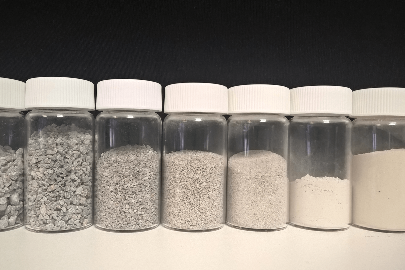Seven clear sample jars displaying limestone materials with progressively smaller particle sizes, ranging from coarse stone fragments to fine powder.