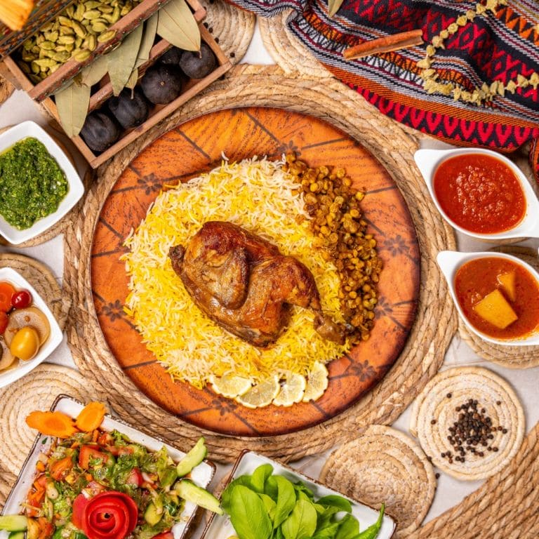 Indian chicken dish on rice on a big orange plate; photo taken from above. Dish surrounded by many other colorful dishes containing sauces, side dishes, toppings